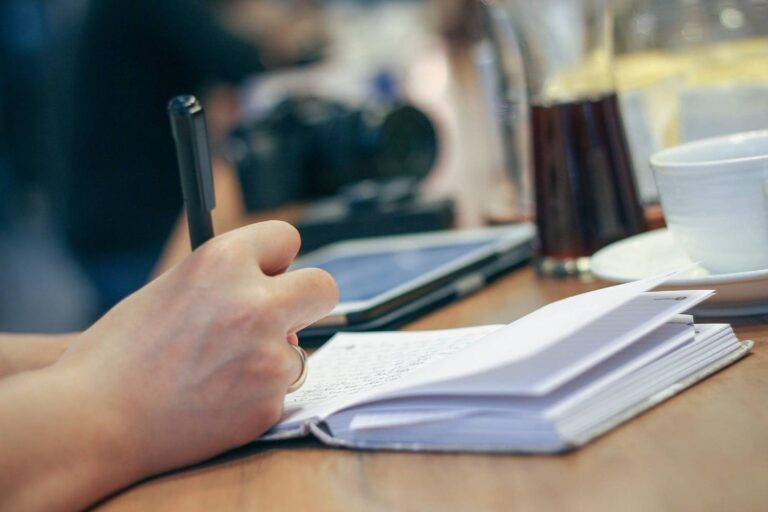 pessoa com uma caneta na mão escrevendo em um livro que está em cima de uma mesa de madeira
