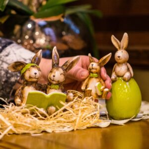 Decoração feita com produtos para Páscoa, como ninho de ovos coloridos, coelhinhos e galhos secos sobre uma mesa de madeira clara, com uma parede de tijolos brancos no fundo.